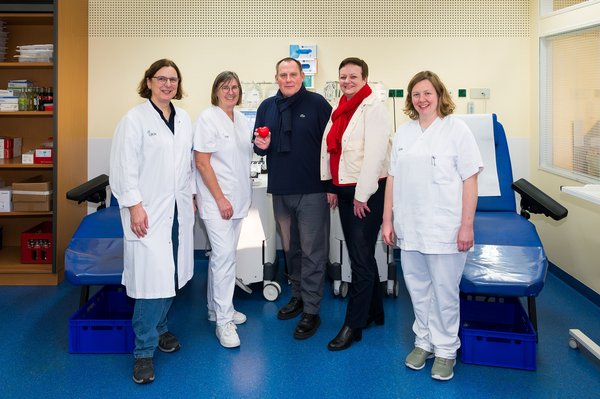 UKM | Ein Ehepaar und drei Mitarbeiterinnen in der UKM-Blutspende. Der Mann hält ein rotes Herz in der Hand. Zwei Liegen im Hintergrund